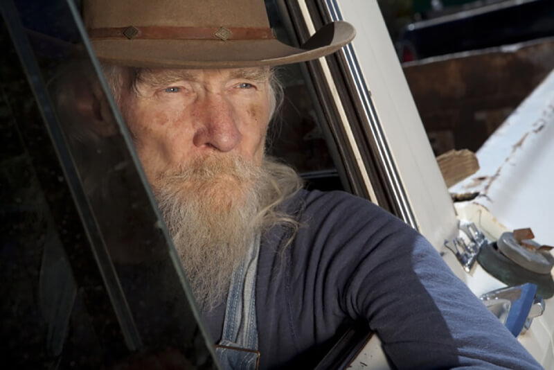Thoughtful older veteran with long white beard and cowboy hat looking out a window, representing the story of overcoming joint pain from military service